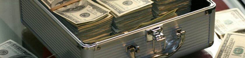 Open briefcase filled with stacks of hundred dollar bills on a glass table, representing wealth.